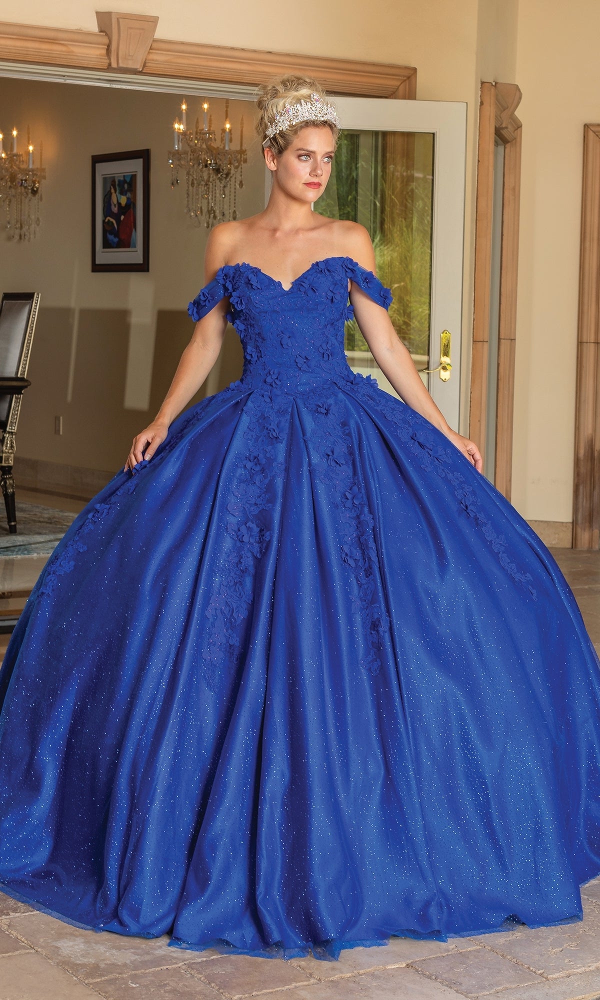 A woman wearing the Quinceanera Dress 1790 By Dancing Queen, a royal blue off-the-shoulder ball gown with floral details and a tiara, stands indoors near an open door.